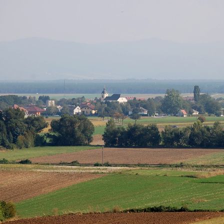 Eine ländliche Landschaft mit einer Ansammlung von Häusern, einige mit roten Dächern, und einer Kirche mit einem Turm. Das Gebiet ist von Feldern und Bäumen umgeben, mit Bergen in der Ferne unter einem vernebelten Himmel.