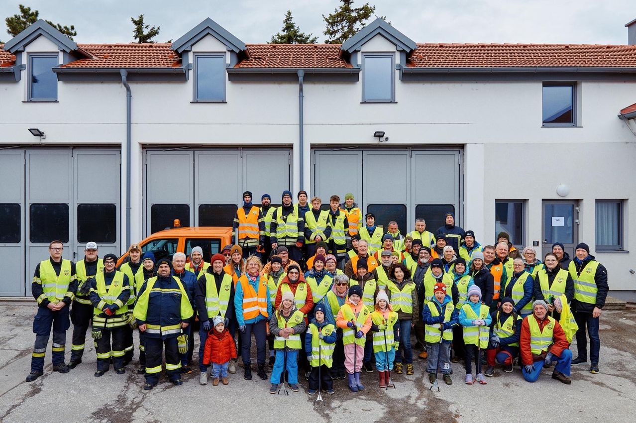 Eine große Gruppe von Menschen, darunter Erwachsene und Kinder, die reflektierende Westen tragen und Stöcke halten, posiert vor einem Gebäude mit zwei Garagen und einem geparkten Fahrzeug davor.