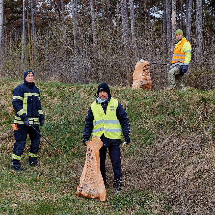 Drei Personen säubern einen Bereich und halten Müllsäcke. Einer hat Besen und Kehrschaufel. Sie tragen reflektierende Westen.