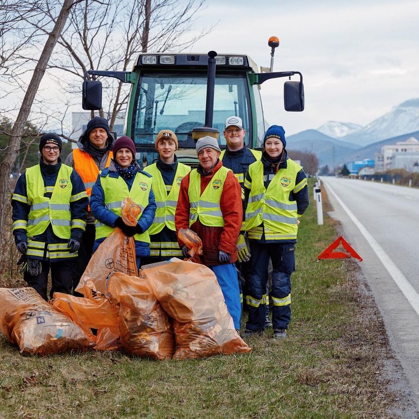 Eine Gruppe von Menschen in gelben Westen posiert neben einem Traktor und Säcken mit Müll am Straßenrand. Dahinter sind Berge zu sehen.