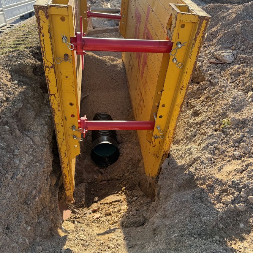 Ein gelbes Baugerät mit roten Stäben und einem schwarzen Rohr ist im Boden vergraben. Der Boden ist mit Sand und Steinen bedeckt.