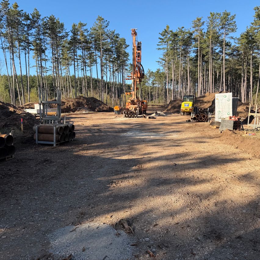 Baustelle mit schweren Maschinen in einem bewaldeten Gebiet. Eine Bohrmaschine ist in Betrieb, mit Baumstämmen und Rohren in der Nähe.