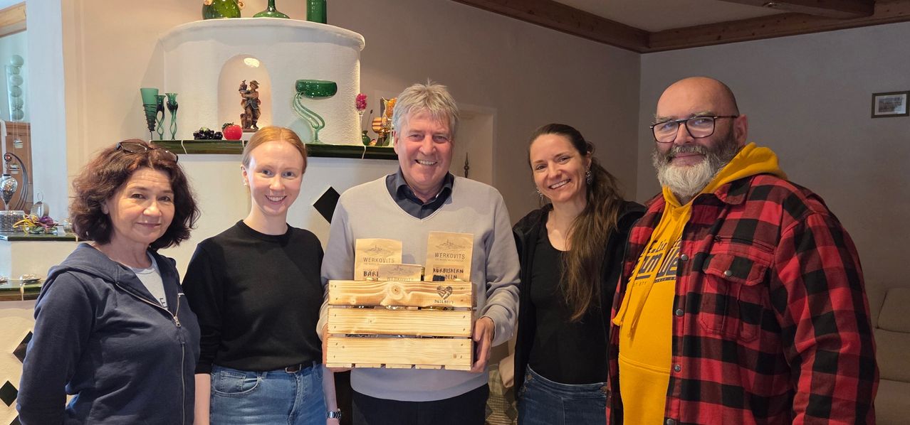 Vier Personen sind drinnen, lächeln und posieren für ein Foto. Ein Mann hält eine Holzkiste mit Produkten. Der Raum hat einen Kamin und verschiedene Dekorationsartikel.