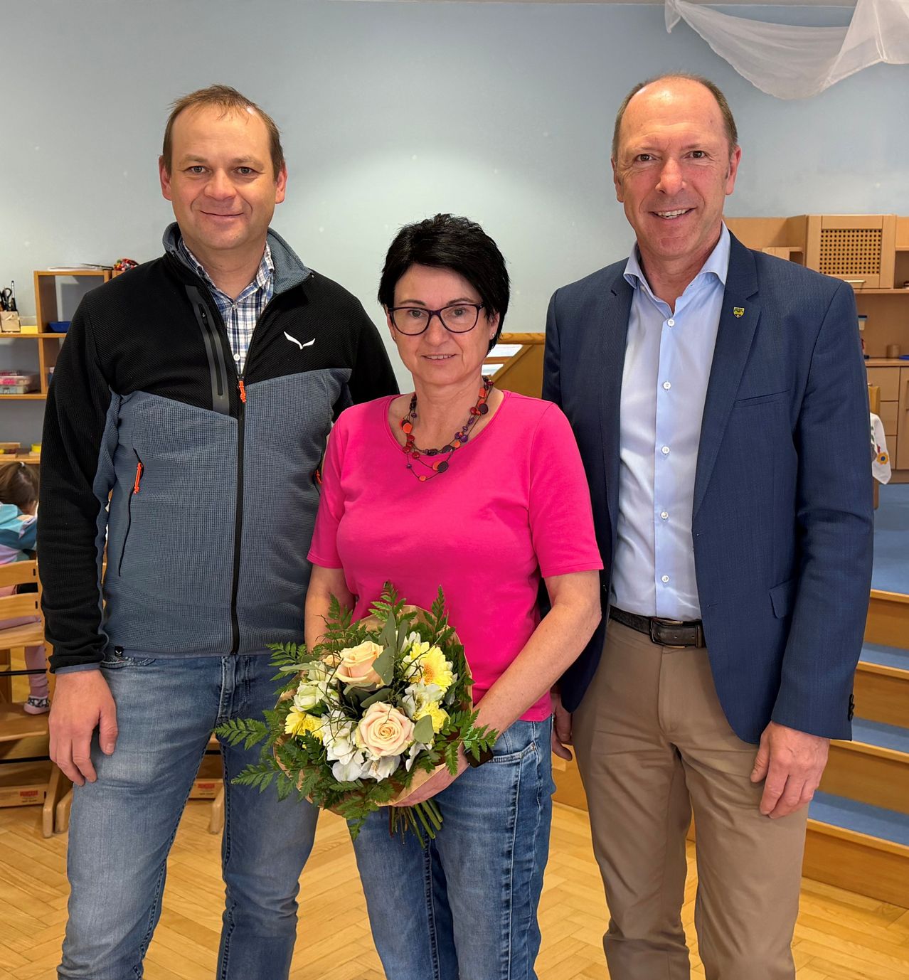Drei Personen, zwei Männer und eine Frau, stehen lächelnd für ein Foto zusammen. Die Frau hält einen Blumenstrauß. Dahinter befindet sich ein Holzregal und eine Treppe.