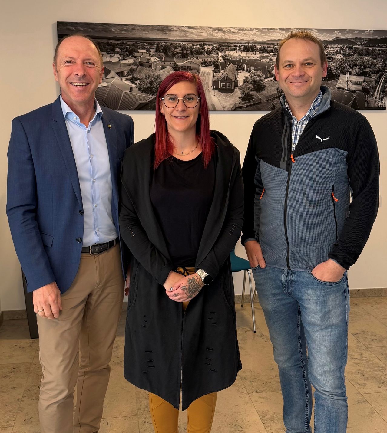 Drei Personen stehen lächelnd in einem Büro. Ein Mann im blauen Blazer, eine Frau mit roten Haaren und ein Mann im grau-schwarzen Mantel. Hinter ihnen ist ein schwarz-weißes Landschaftsfoto.