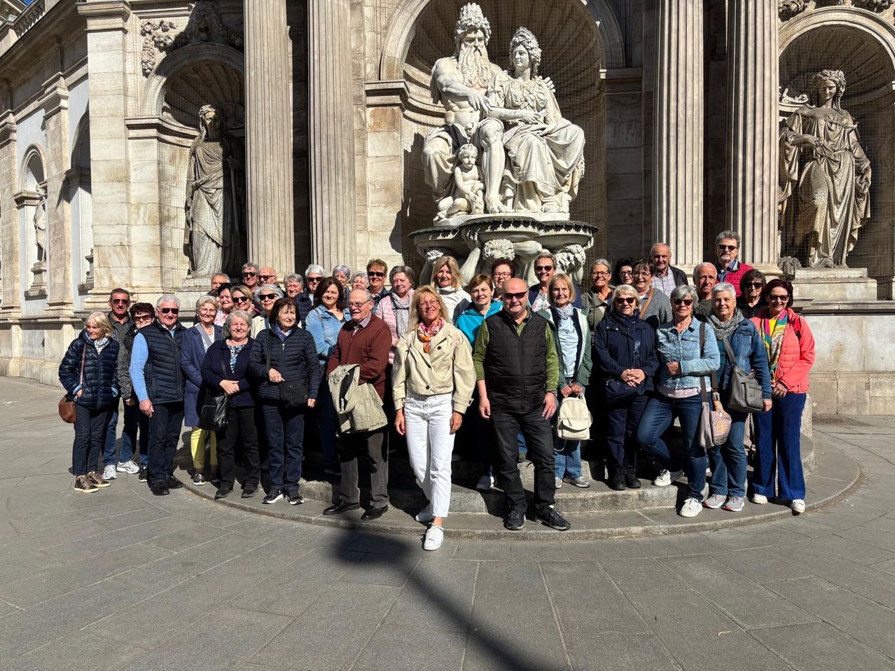 Eine Gruppe von Menschen steht vor einem großen Brunnen in einer historischen Umgebung, lächelt und posiert für ein Foto.