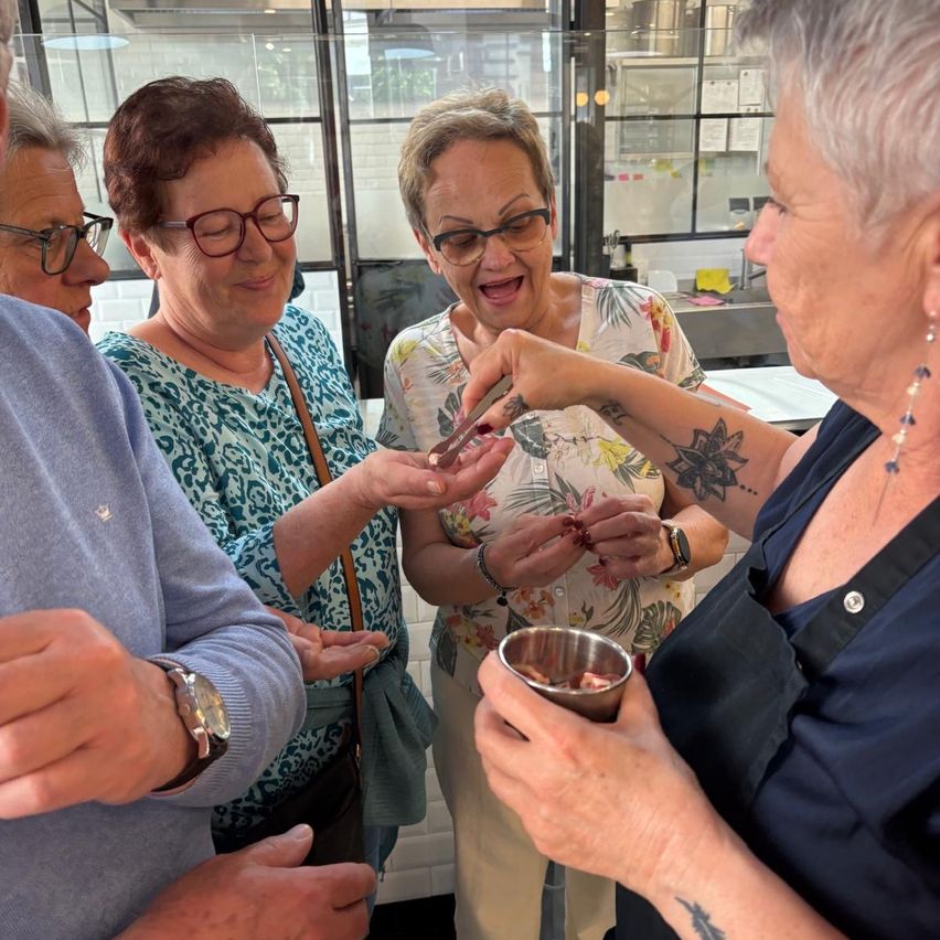 Eine Gruppe älterer Frauen in einer Küche, eine mit einer schwarzen Schürze, hält einen kleinen Teller mit Essen und ein Stück Papier und schaut sich etwas vor ihnen an.