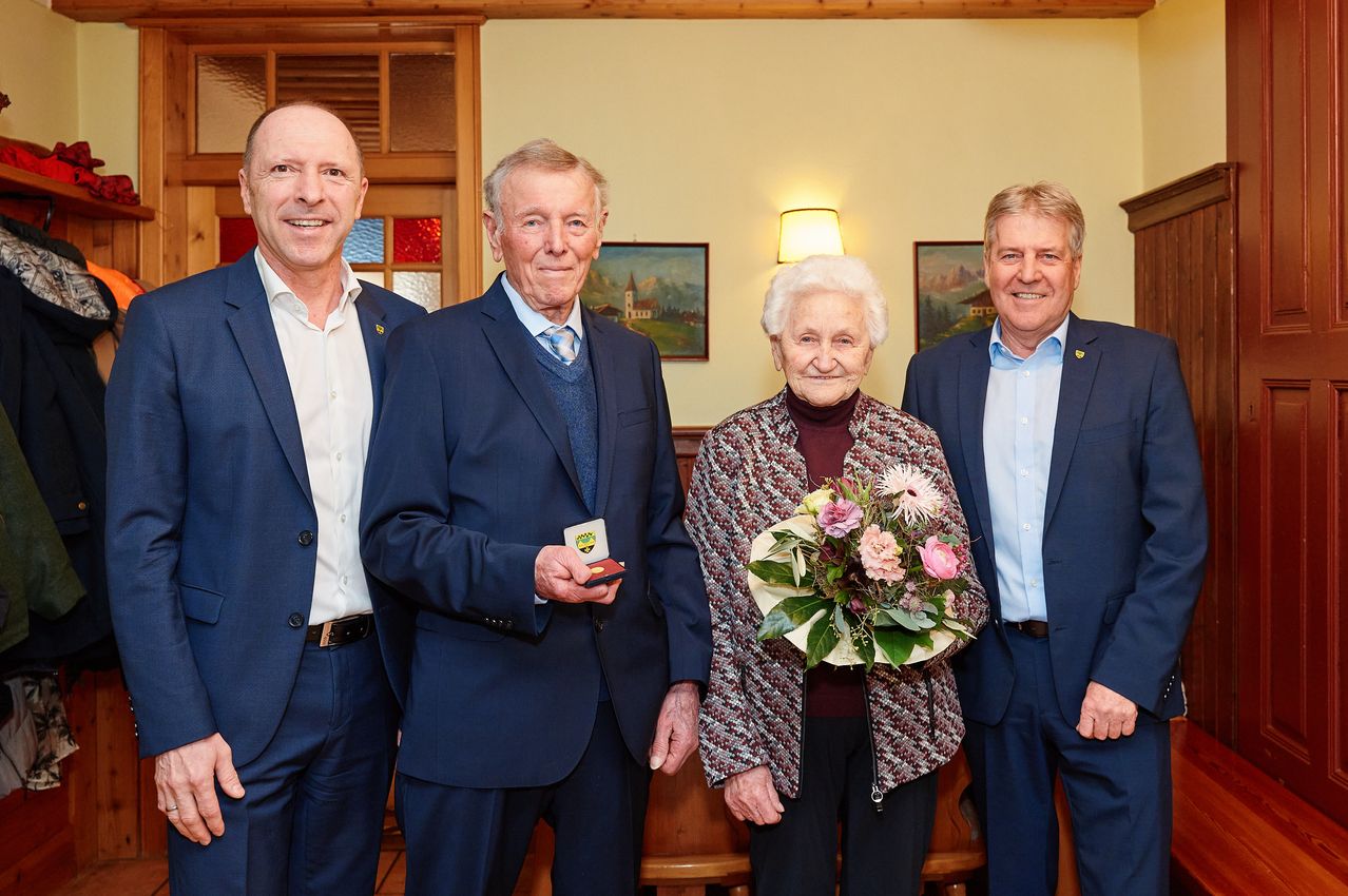 Four adults stand in a room, smiling. The elderly woman holds a bouquet of flowers. The man next to her holds a medal. The other two men wear suits.