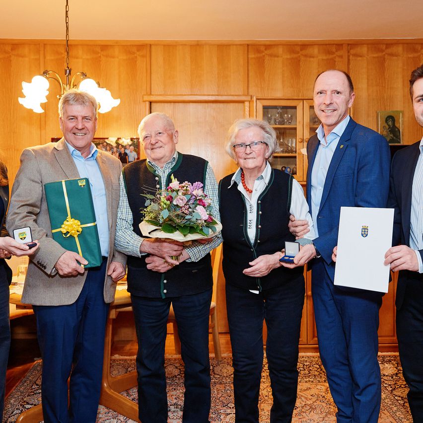 Fünf Personen stehen in einem Raum und lächeln für ein Foto. Sie halten Blumen, Urkunden und Geschenke. Holzwände und eine Lampe sind im Hintergrund.