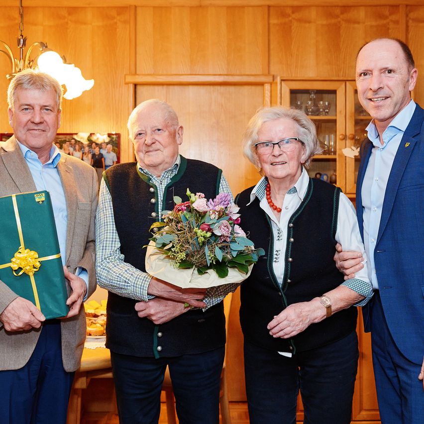 Vier Erwachsene stehen zusammen in einem Raum und lächeln für ein Foto. Ein Mann auf der linken Seite hält ein verpacktes Geschenk. Der Mann in der Mitte hält einen Blumenstrauß.