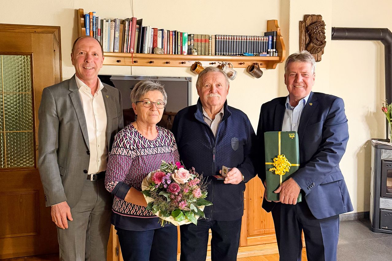 Vier Personen stehen in einem Raum. Der Mann auf der linken Seite trägt einen Anzug und einen Gürtel. Die Frau hält einen Blumenstrauß. Der Mann in der Mitte hält eine Karte. Der Mann auf der rechten Seite hält eine Geschenkbox.