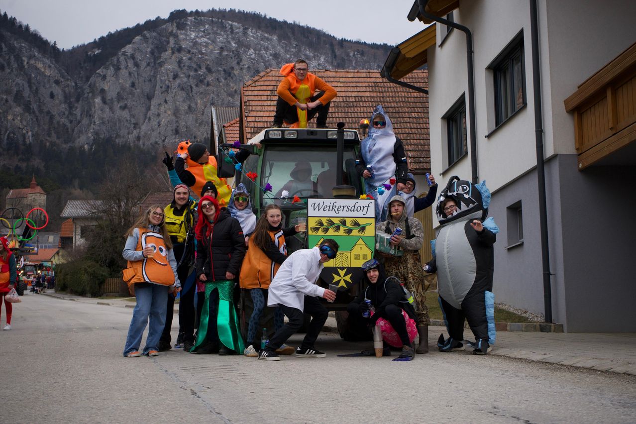 Eine Gruppe von Menschen in verschiedenen Kostümen posiert für ein Foto vor einem Traktor mit dem Schild Weikersdorf. Ein Berg ist im Hintergrund.
