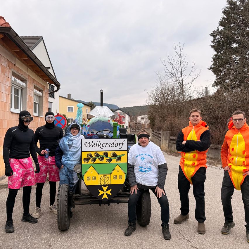 Eine Gruppe von Menschen in Kostümen und mit Schutzbrillen steht vor einem geschmückten Fahrzeug mit dem Ortsnamen Weikersdorf. Sie sind draußen, mit Häusern und Bäumen im Hintergrund.