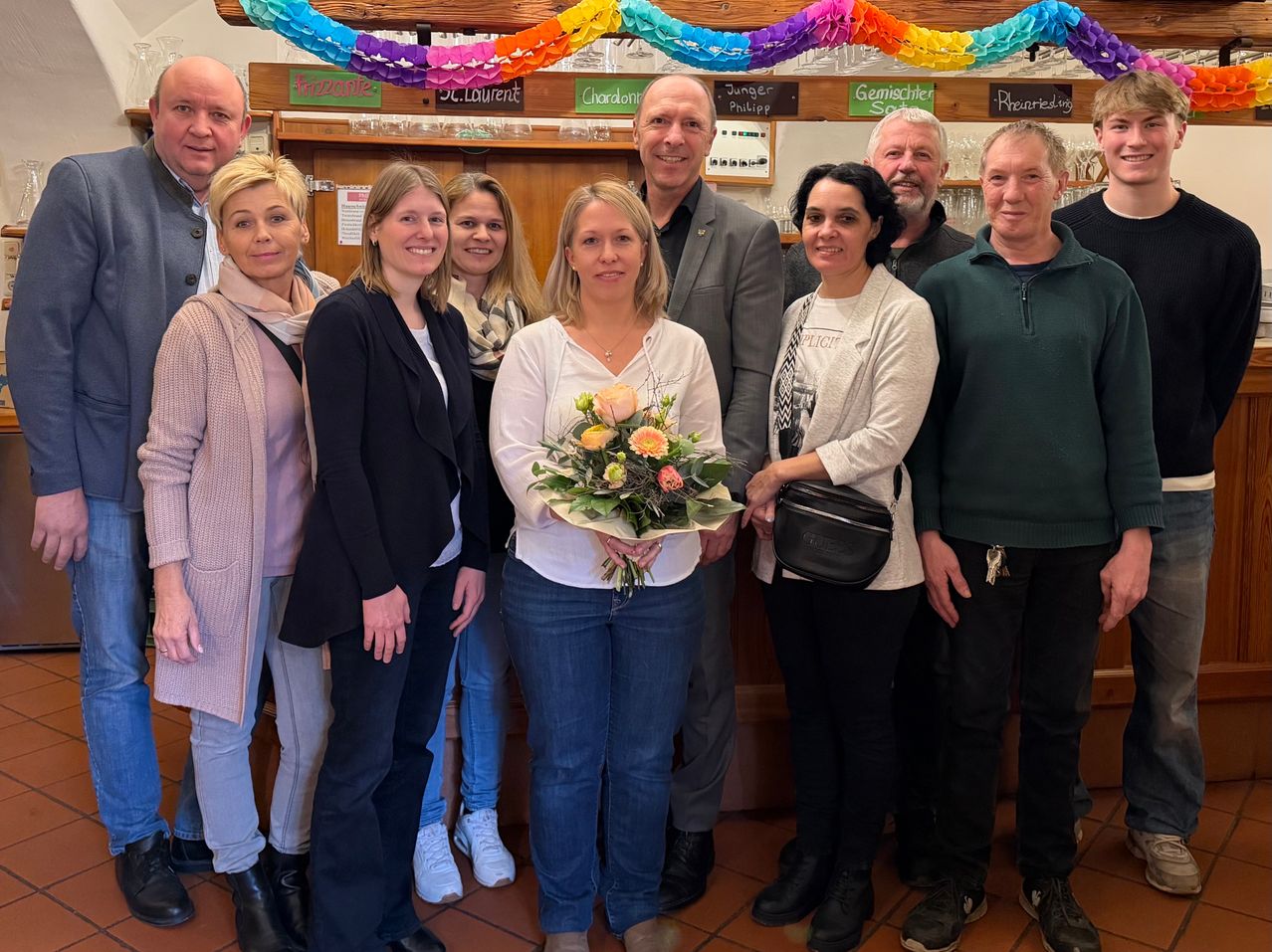 Eine Gruppe von Erwachsenen steht in einem Raum und lächelt. Eine Frau in der Mitte hält einen Blumenstrauß. Hinter ihnen ist ein Holzregal mit verschiedenen Getränken zu sehen. Banner mit bunten Papierblumen hängen darüber.