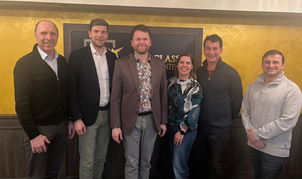 Fünf Personen stehen zusammen vor einem Schild. Das Schild hat schwarze und gelbe Farben. Sie alle lächeln für die Kamera.