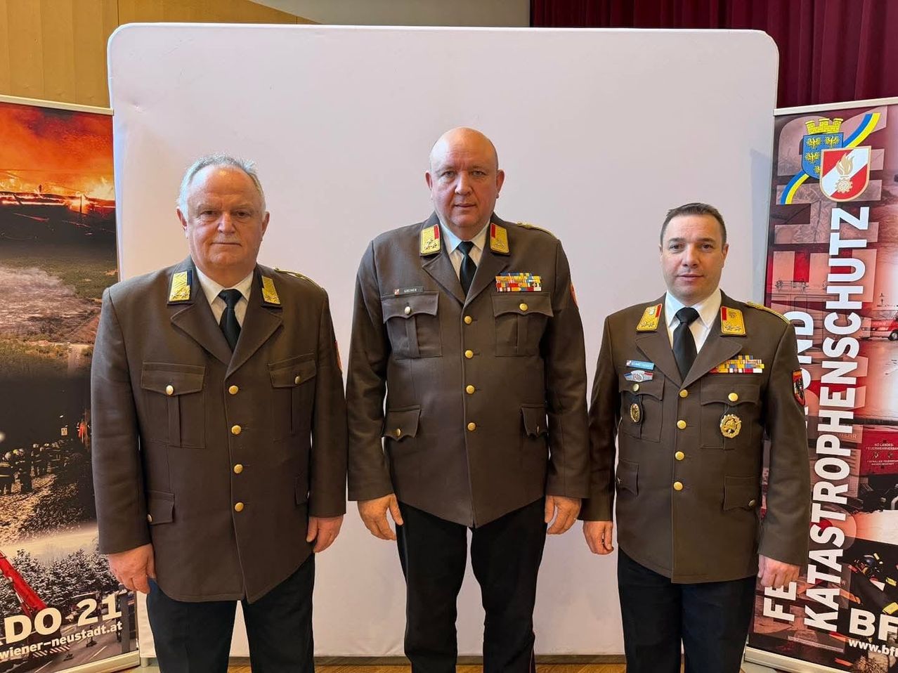 Drei Männer in Militäruniformen stehen nebeneinander vor einem weißen Hintergrund. Der Mann in der Mitte ist kahlköpfig und trägt eine Krawatte.