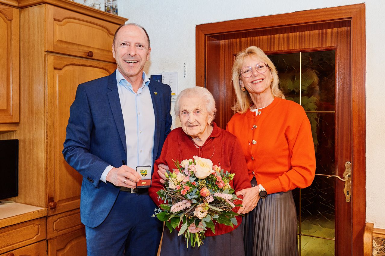 Drei Menschen stehen lächelnd drinnen zusammen. Der Mann auf der linken Seite hält eine Medaille. Die ältere Frau in der Mitte hält einen Blumenstrauß. Die Frau auf der rechten Seite trägt eine Brille und einen orangefarbenen Pullover.