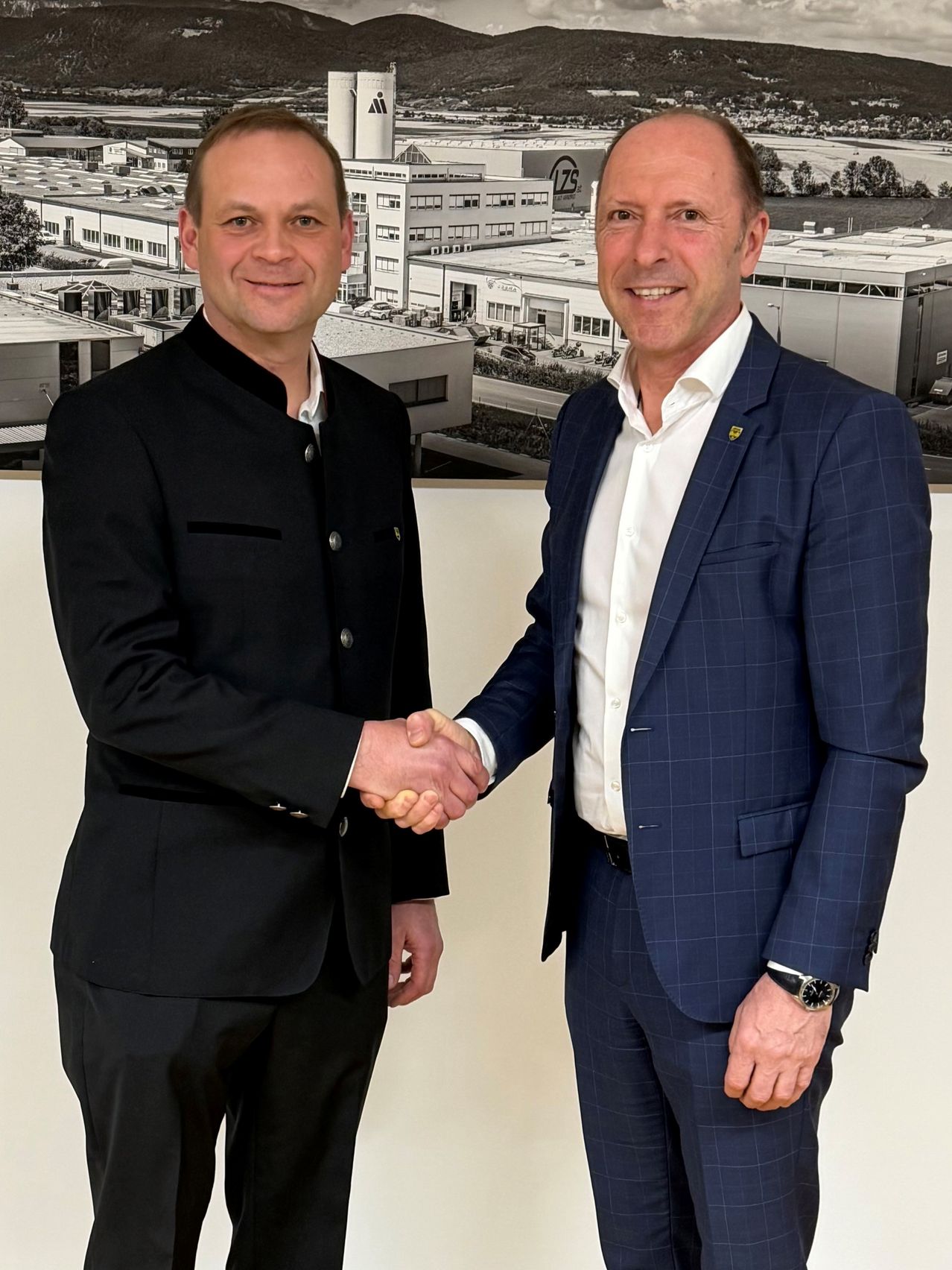 Zwei Männer stehen und schütteln sich die Hände vor einem großen Foto eines Industriegebäudes. Beide Männer tragen formelle Anzüge.