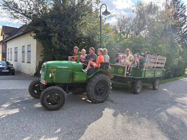 Eine Gruppe von Menschen fährt auf einem grünen Traktor mit Anhänger auf einer Straße, mit einem Haus und Bäumen im Hintergrund.