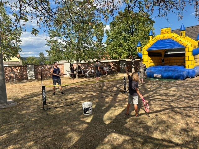 Eine Gruppe von Menschen spielt an einem sonnigen Tag Badminton im Garten mit einem aufblasbaren Schloss im Hintergrund.