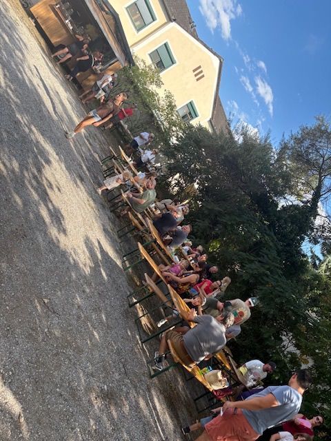 Eine Gruppe von Menschen sitzt an Tischen vor einem Gebäude an einem sonnigen Tag. Die Tische sind in Reihen angeordnet, und einige Menschen stehen in der Nähe der Tische. Das Gebäude hat eine gelbe Wand mit Fenstern und einer Tür. Bäume und Pflanzen stehen vor dem Gebäude.