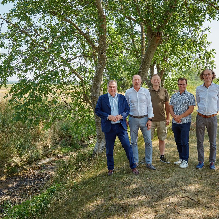 Fünf Männer stehen in einer Reihe auf einem Feld, mit Bäumen im Hintergrund. Sie sind in formeller und lässiger Kleidung gekleidet.