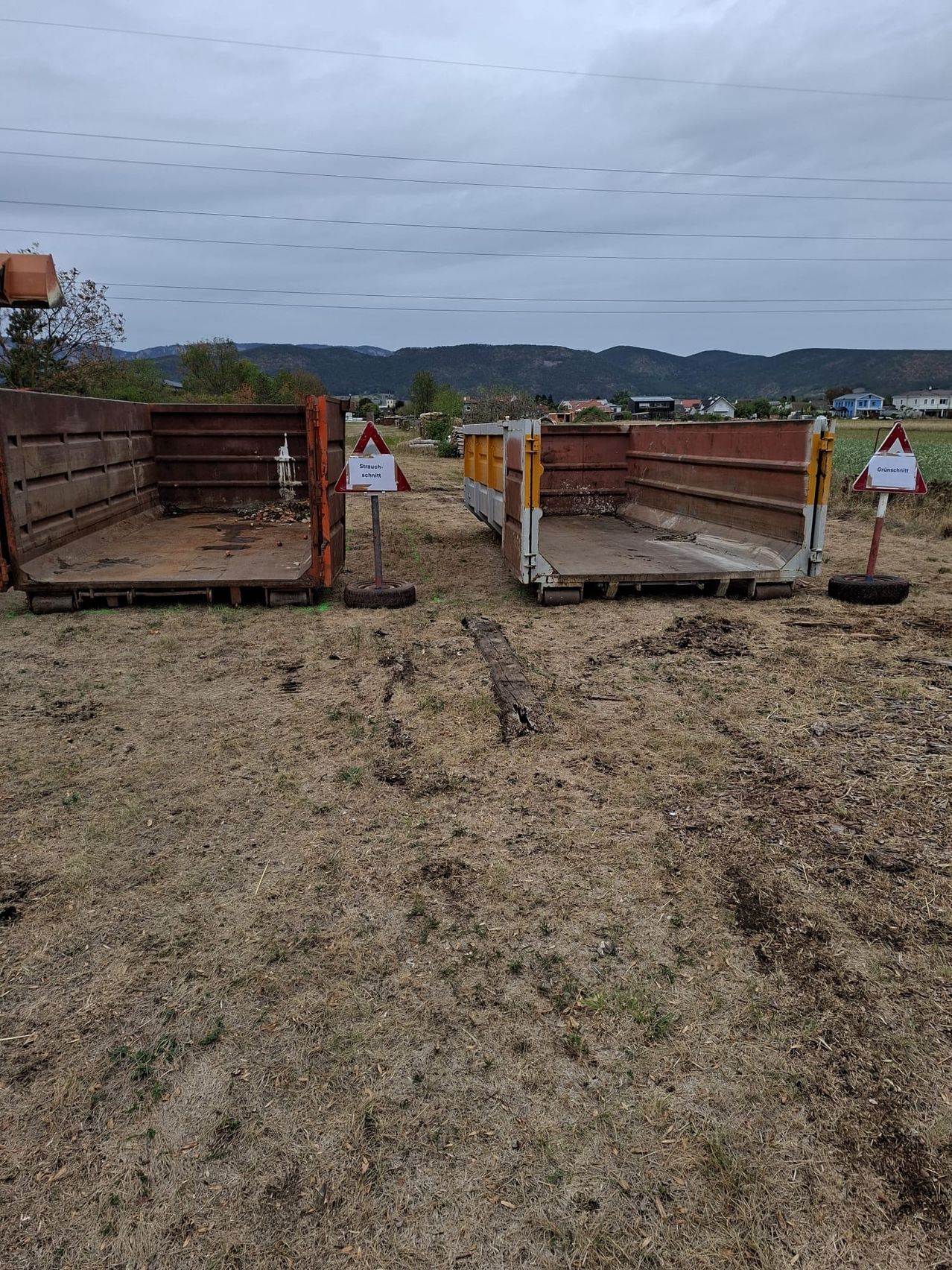 Zwei offene Müllcontainer stehen auf einem Feld mit Bergen im Hintergrund. Ein Container hat ein Schild mit der Aufschrift 'Straach'.
