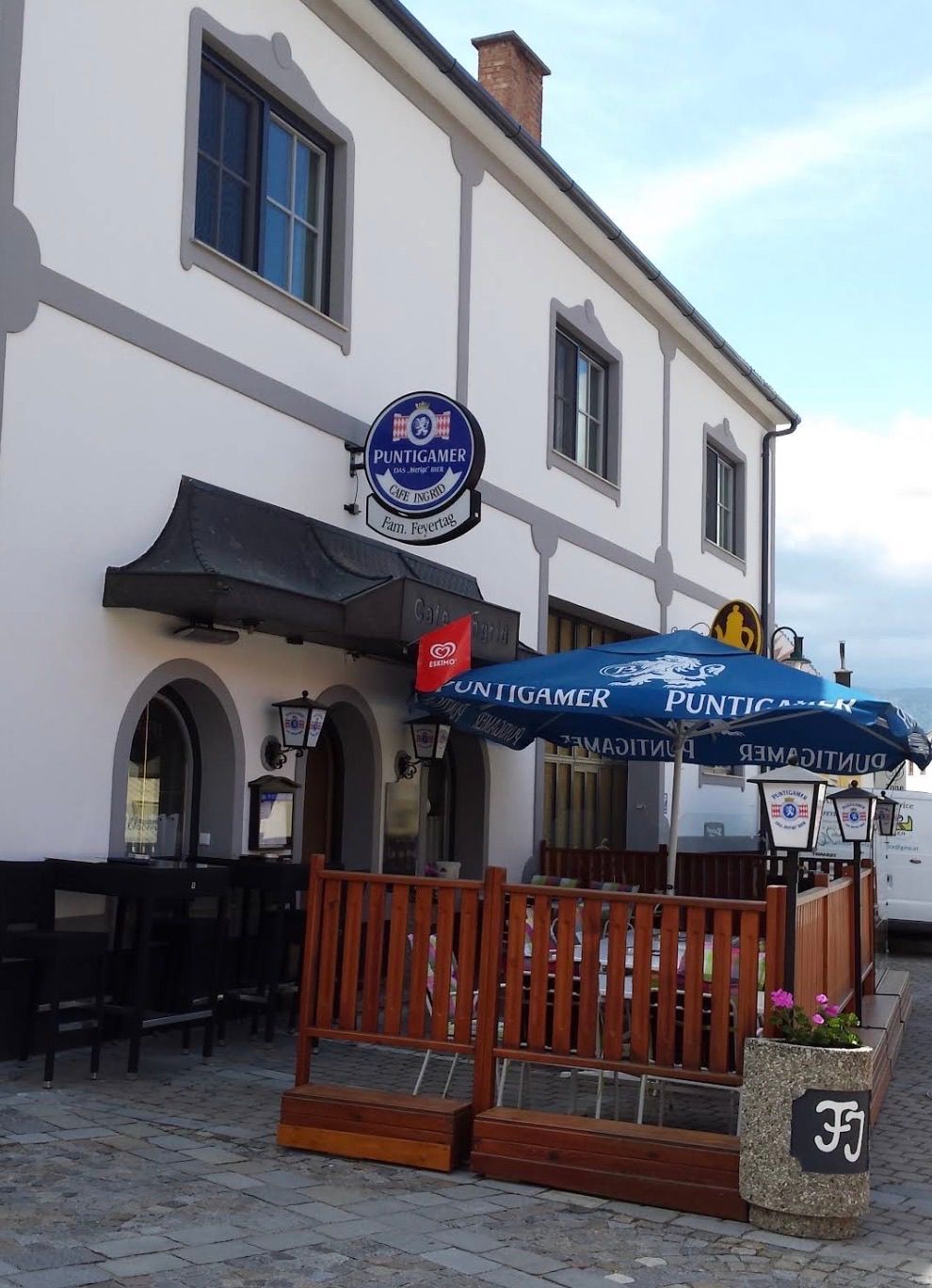Ein Café mit einem blauen Regenschirm und einem Schild, das Puntigamer Cafe in Erd anzeigt. Das Gebäude hat bogenförmige Fenster und einen schwarzen Vorbau.