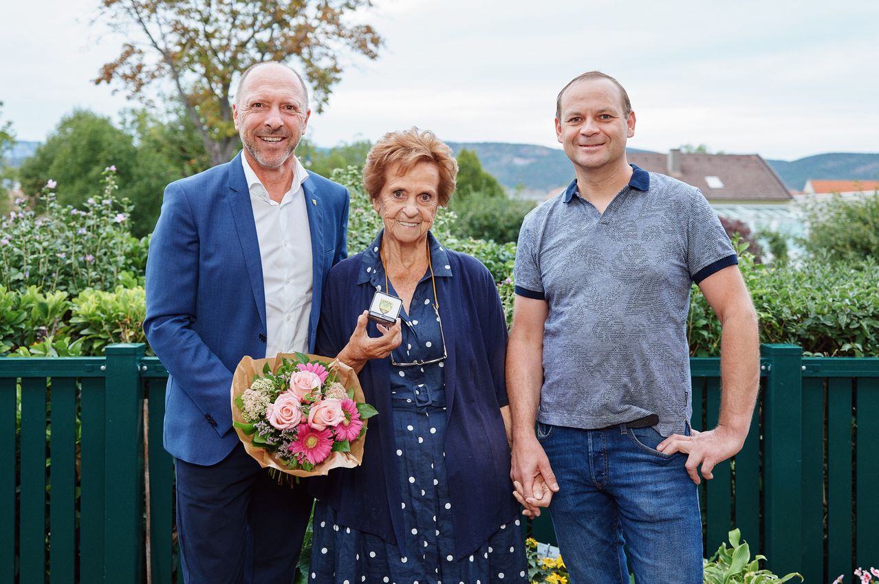 Eine ältere Frau hält eine kleine Trophäe und einen Blumenstrauß und steht zwischen zwei lächelnden Männern in einem Garten.