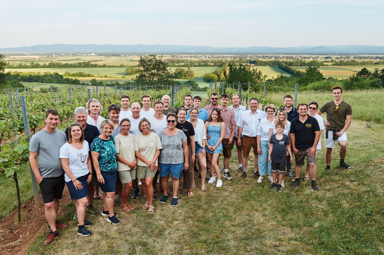 Eine Gruppe von Menschen in Freizeitkleidung posiert für ein Foto in einem Weinberg mit einer malerischen Aussicht auf die Landschaft.