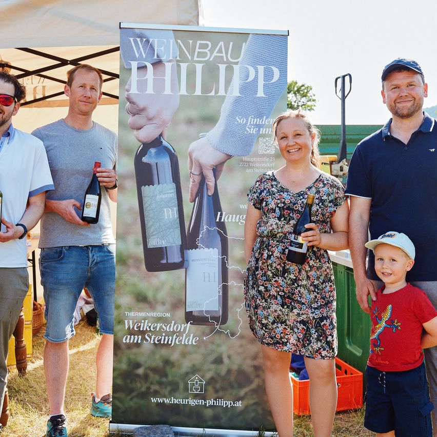 Fünf Menschen, darunter eine Frau und ein kleines Kind, lächeln und halten Weinflaschen. Sie stehen vor einem großen Banner mit dem Text 'Weinbau Philipp' und einer Karte.