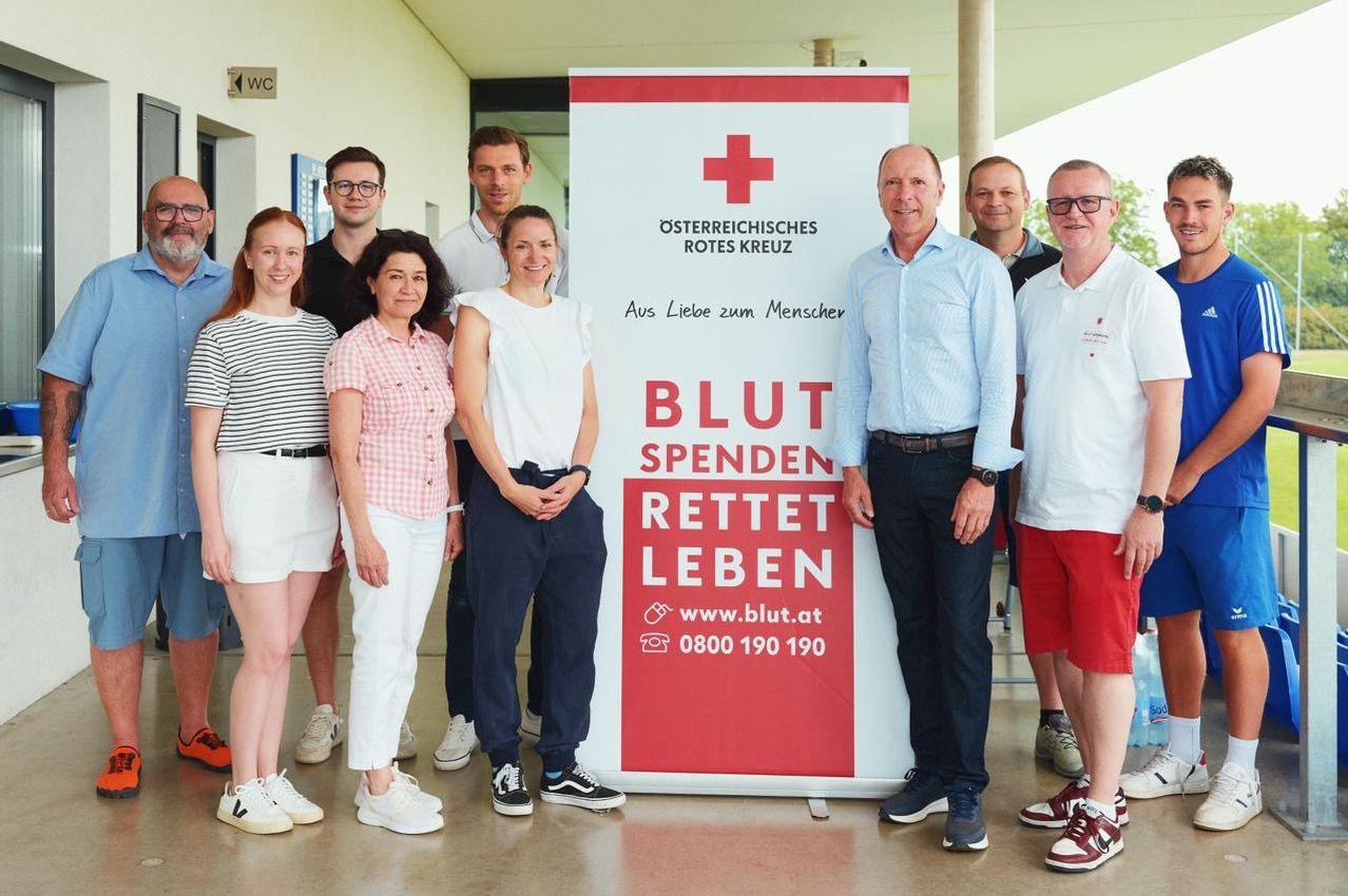 Eine Gruppe von Menschen steht vor einem Banner. Es steht 'Blut Spendet Rettet Leben' und hat eine Website und Telefonnummer. Die Menschen lächeln und scheinen für ein Foto zu posieren.
