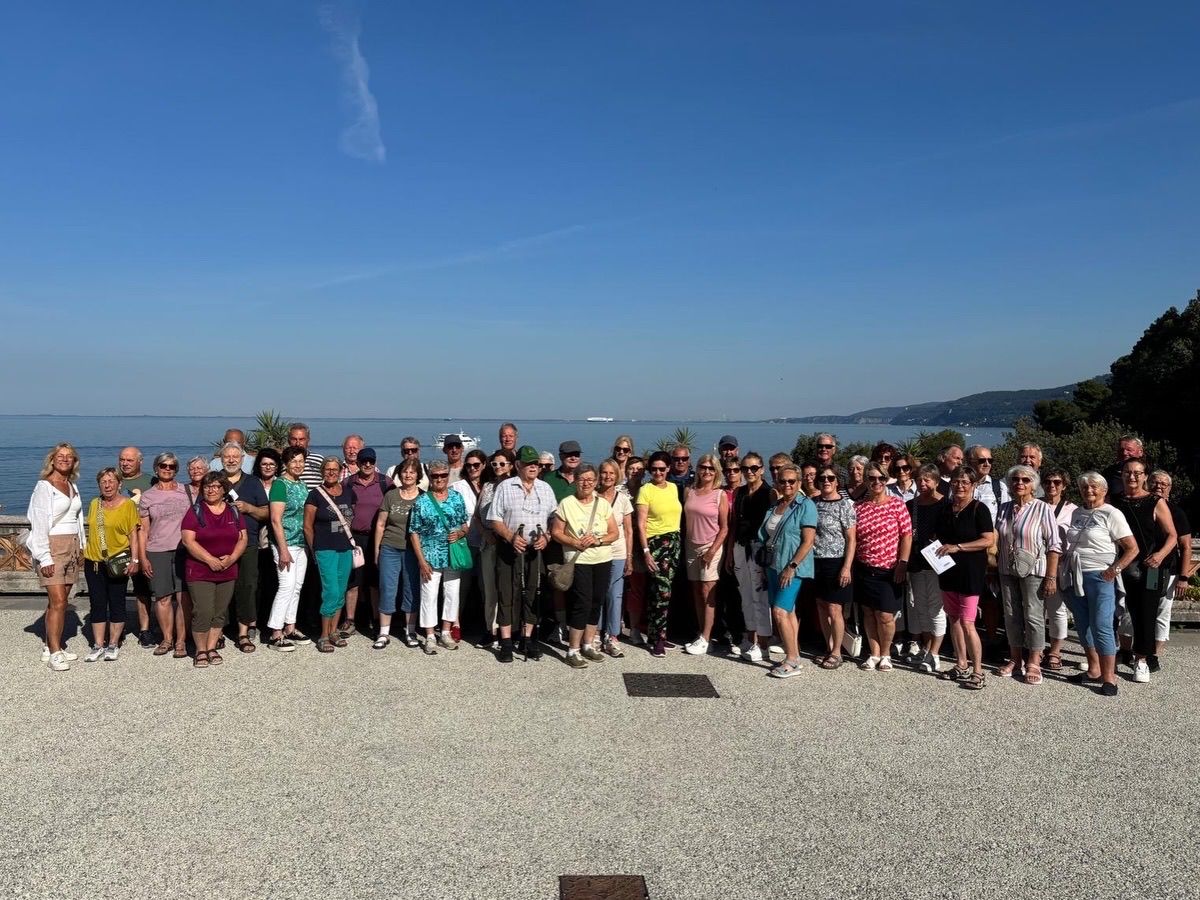 Eine Gruppe von Menschen steht in einer Reihe zusammen, wahrscheinlich am Strand, mit einem ruhigen Ozean im Hintergrund. Sie alle lächeln und scheinen für ein Foto zu posieren.