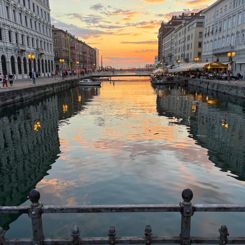 Bei Sonnenuntergang ist der Canal Grande ruhig und reflektiert Orange- und Rosatöne. Gebäude säumen beide Seiten, und Menschen schlendern auf dem Bürgersteig. Boote sind festgemacht, und der Himmel ist mit Wolken bemalt.