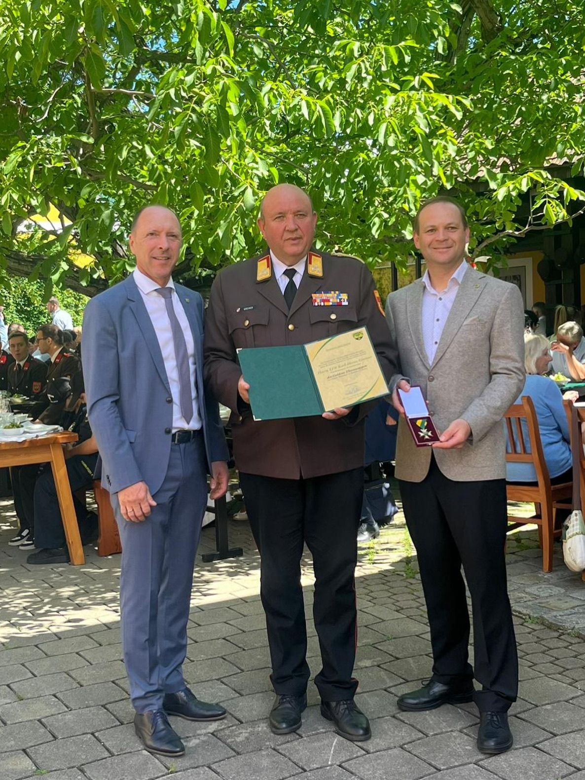 Drei Männer stehen in einem Außenbereich. Der zentrale Mann in Uniform hält ein Zertifikat und eine Medaille, während die Männer auf beiden Seiten Anzüge und Krawatten tragen. Dahinter sitzen Menschen an Tischen, einige in Uniformen.