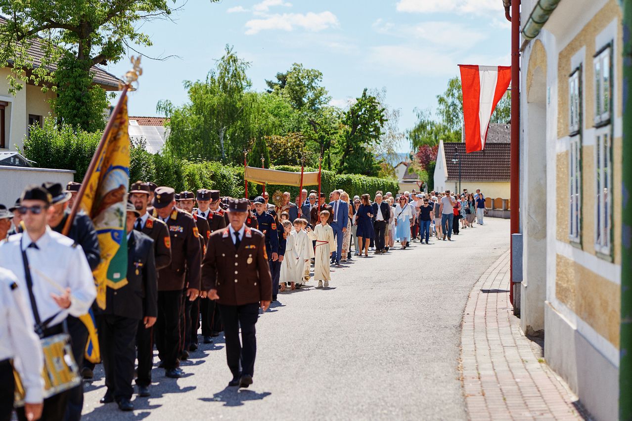 Eine Prozession von Personen in Uniformen und Kindern in weißen Gewändern geht eine Straße hinunter, an der Seitenflaggen wehen.