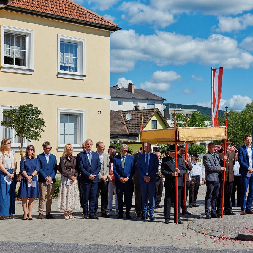 Eine Gruppe formell gekleideter Menschen steht vor einem Gebäude mit einer rot-weißen Fahne unter einem wolkenreichen Himmel.