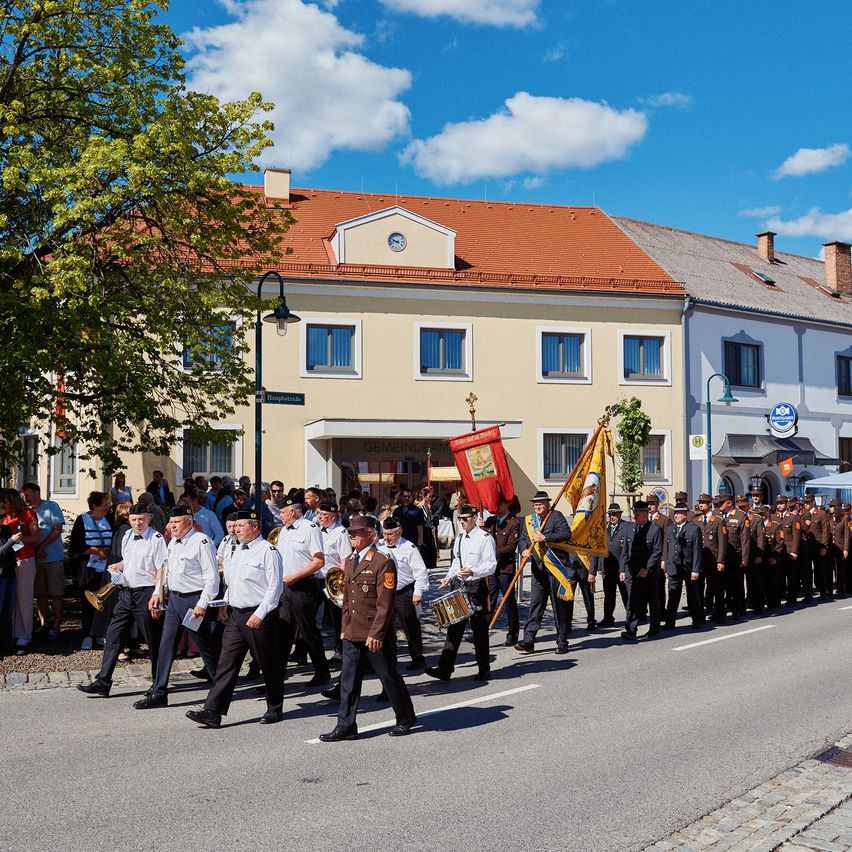 Ein Marsch von Militärpersonal auf der Straße, mit Flaggen und Trommeln, vor einem Gebäude mit roten Dächern und einem Baum.