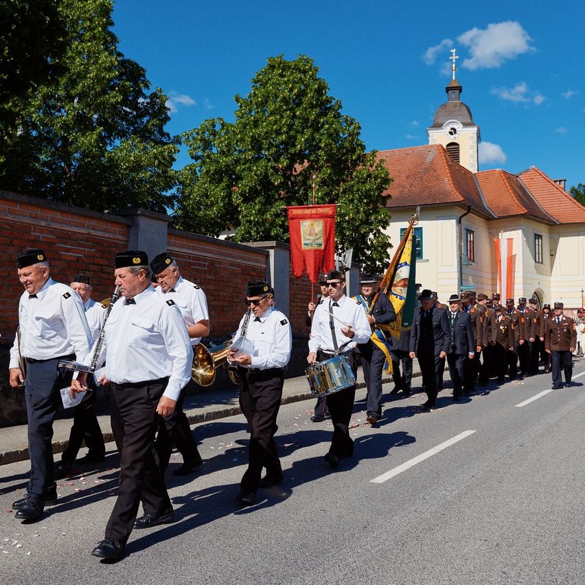 Eine Musikkapelle spielt Instrumente und trägt Flaggen während einer Parade vor einem Kirchengebäude unter blauem Himmel.