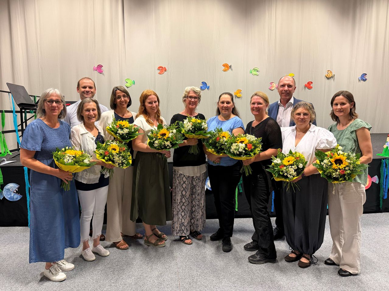 Eine Gruppe von Menschen steht zusammen und hält Blumensträuße. Sie lächeln und scheinen für ein Foto zu posieren. Der Hintergrund zeigt einen weißen Vorhang, der mit farbenfrohen Fischausschnitten verziert ist.