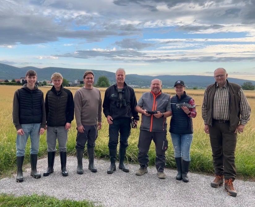 Sieben Personen stehen in einem Feld mit Bergen im Hintergrund, tragen Stiefel und halten teilweise Fernsteuerungen. Sie lächeln und scheinen für ein Foto zu posieren.
