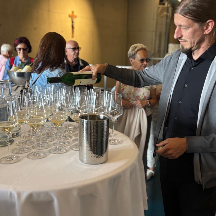 Ein Mann schenkt Wein in Gläser auf einem Tisch, umringt von zuschauenden Menschen. Ein Kreuz ist an der Wand sichtbar.