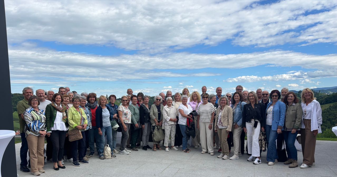Eine große Gruppe von Menschen, überwiegend Frauen, versammelt sich an einem sonnigen Tag und posiert für ein Foto. Sie sind lässig gekleidet. Der Hintergrund zeigt eine Landschaft mit einem bewölkten Himmel.