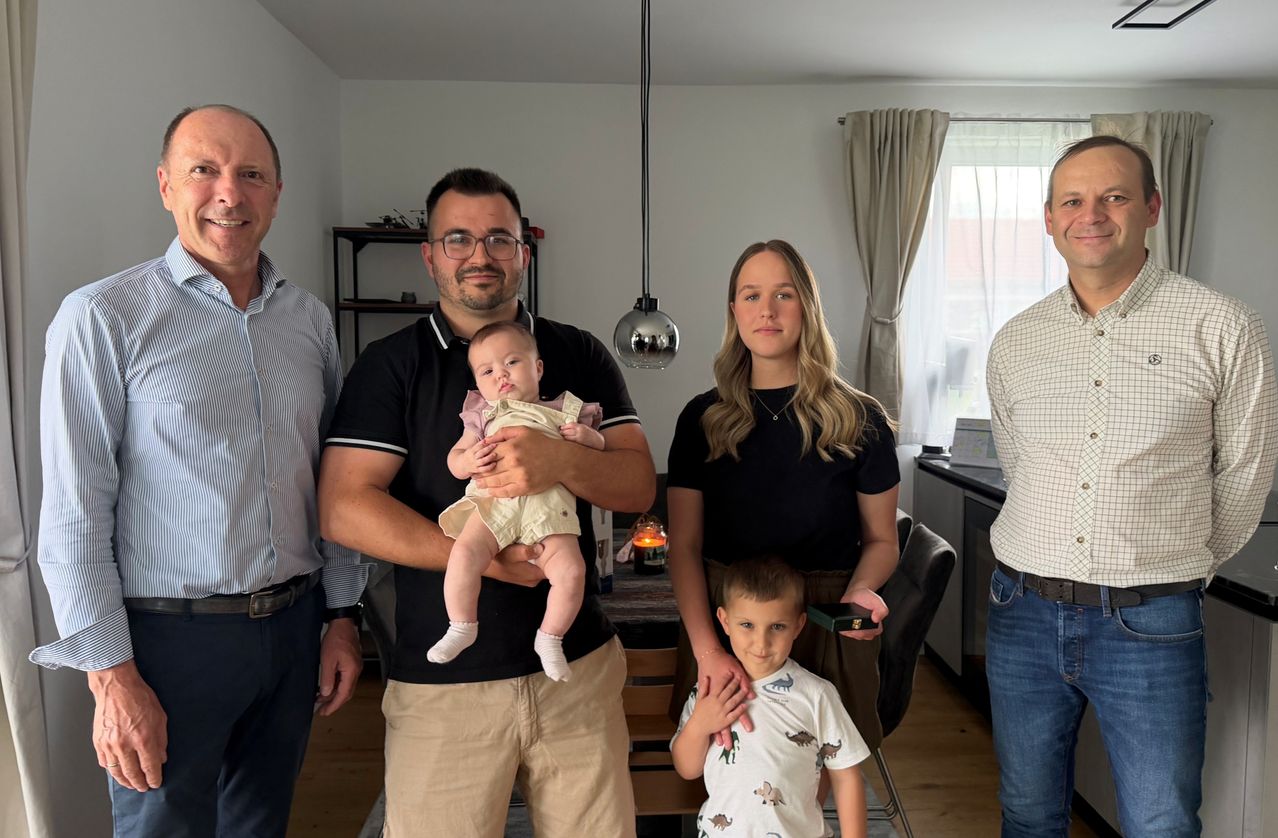 Fünf Personen stehen in einem Raum. Ein Mann hält ein Baby und eine Frau ein Kind. Ein anderer Mann steht links. Eine Lampe hängt von der Decke.