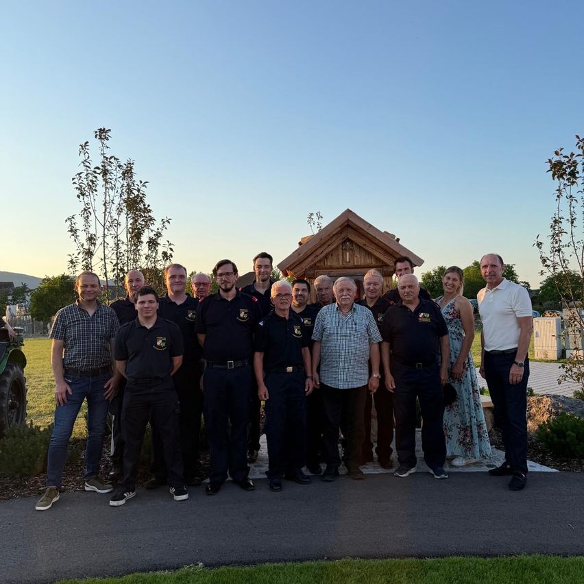 Eine Gruppe von Menschen steht vor einem kleinen Holzhaus. Einige tragen Uniformen, andere Freizeitkleidung. Hinter ihnen sind Pflanzen und ein Traktor. Der Himmel ist blau und klar.