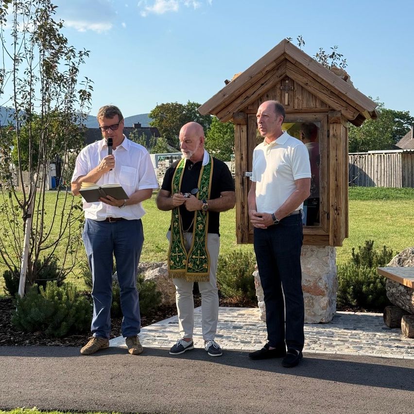 Drei Männer stehen bei einer Holzkapelle, einer hält ein Mikrofon und ein Buch. Ein anderer trägt eine grüne Stola. Sie stehen auf einem gepflasterten Weg in einem Park.