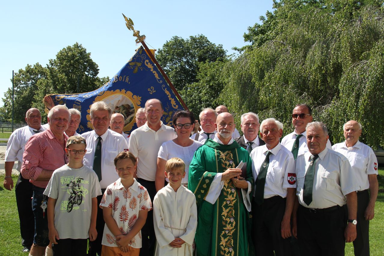 Eine Gruppe von Menschen in formeller Kleidung steht für ein Foto in einem Park. Ein Priester in grünen Gewändern steht in der Mitte, mit einem Banner und einem Pfahl hinter ihm.