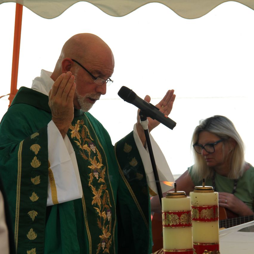 Ein Priester steht mit einem Mikrofon und hält eine Rede, während eine Frau im Hintergrund eine Gitarre spielt.