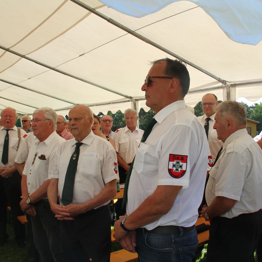 Eine Gruppe von Männern in weißen Hemden und grünen Krawatten steht unter einem Zelt. Der Mann vorne trägt eine Brille und ein weißes Hemd mit einem Abzeichen am Ärmel.