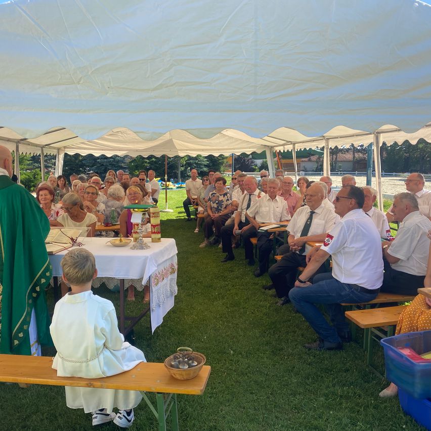 Eine Gruppe von Menschen sitzt auf Bänken unter einem Zelt und hört einem Priester zu. Ein Junge in einer weißen Robe sitzt auf einer Bank vor einem Tisch.